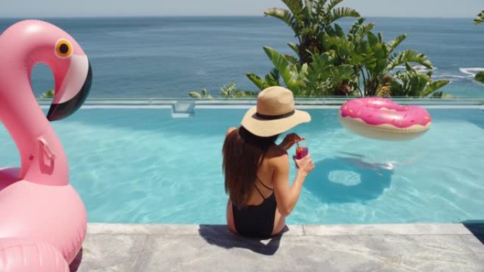 比基尼女人坐在游泳池边喝酒享受夏日晒太阳放松粉色火烈鸟游泳管充气4k