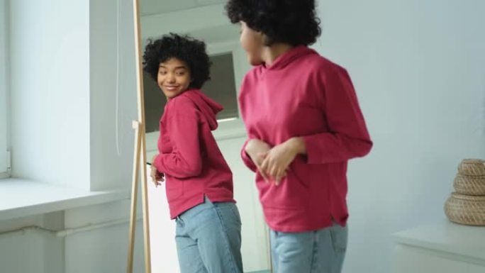 爱自己。美丽的年轻微笑的非洲裔美国妇女跳舞触摸卷发享受她的镜子反射。看着镜子的黑人女士看起来自信快乐