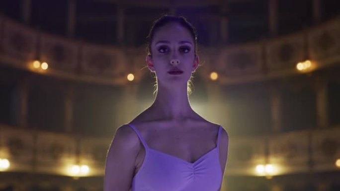 年轻优雅的女性古典芭蕾舞演员在经典剧院里看着镜头，带着戏剧性的灯光微笑的电影镜头。专业女舞蹈表演者自