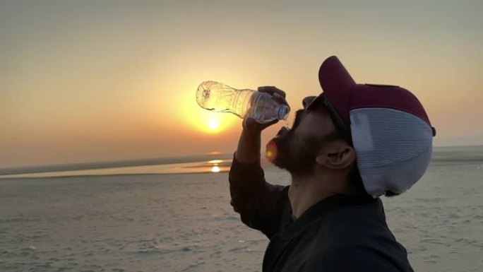 背景是日落，以慢动作的人喝水的特写镜头。戴着易拉罐和太阳镜的男子用slo运动水飞溅取水