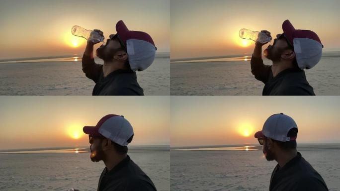 背景是日落，以慢动作的人喝水的特写镜头。戴着易拉罐和太阳镜的男子用slo运动水飞溅取水