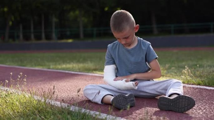 孩子的手臂骨折了。悲伤的白人小男孩断手坐在户外的运动场上。孩子用石膏触摸他骨折的肢体。暑假骨折损伤。