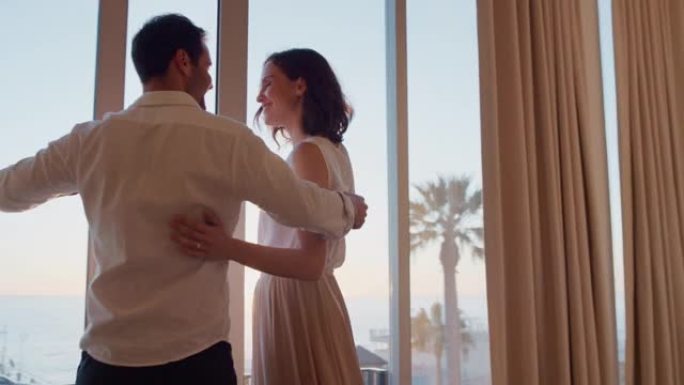 快乐的旅行夫妇在蜜月假期到达酒店房间，看着窗外，享受日落时美丽的海景，拥抱分享浪漫的假期