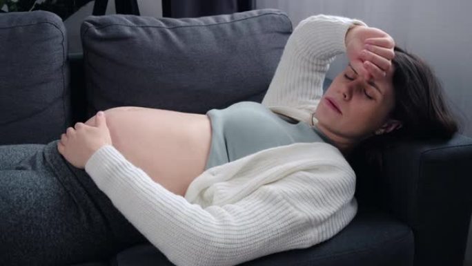对分娩概念感到恐惧。特写疲惫的年轻孕妇患头痛或消极想法，担心不好的产前筛查结果，躺在家里的沙发上