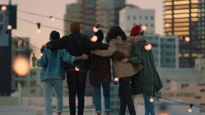 一群快乐的朋友在屋顶上举起手臂庆祝友谊，享受一起喝酒拥抱自由，在日落时看着城市的天际线
