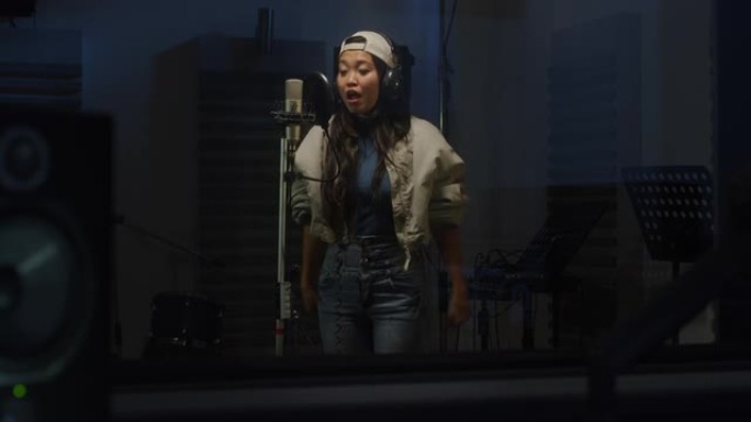 年轻的亚洲女歌手肖像，戴着耳机在专业录音室录制专辑。富有表现力的年轻女子带着情感唱歌，与广播听众分享