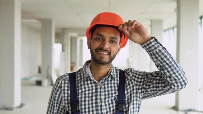 穿着制服的自信的印度亚裔工人建筑商戴着安全帽看着相机，站在建筑工地上