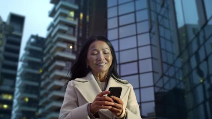 优雅的亚洲中年女商人的肖像在晚上用智能手机从办公室走了出来。女律师检查她的电子邮件，通过社交媒体与家