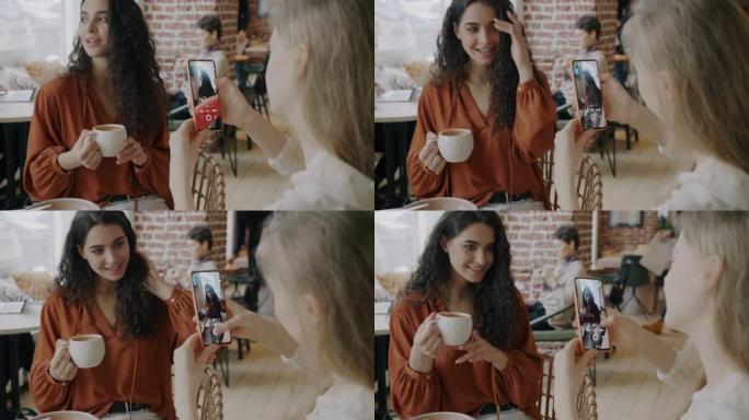 美丽的女孩在咖啡馆里摆姿势，拿着咖啡，朋友用智能手机相机拍照