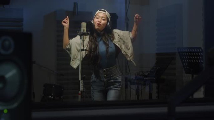 一位专业亚洲女歌手在录音室录制歌曲的肖像。美丽的年轻艺术家喜欢表演她的新单曲，用富有表现力的面孔和手