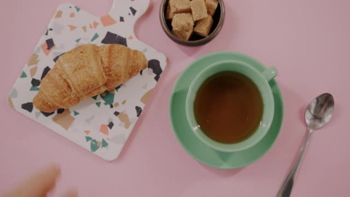 将美味的羊角面包放在粉红色桌子上的茶和糖旁边的手的俯视图