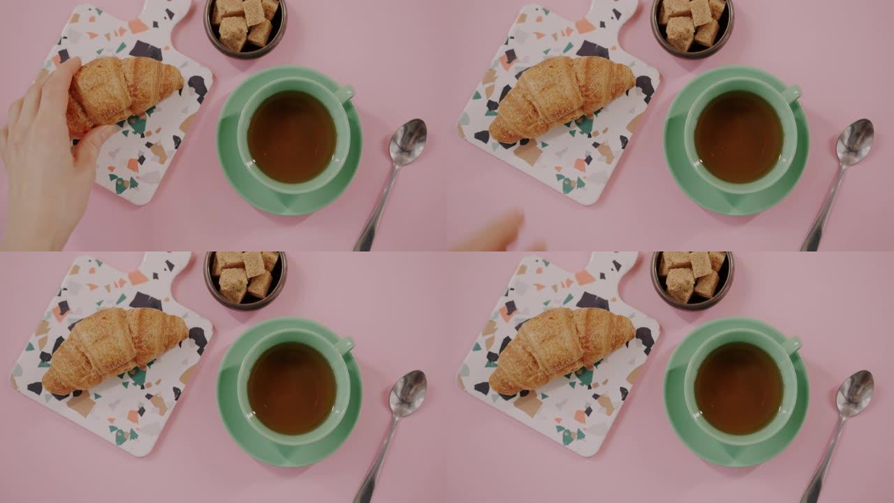 将美味的羊角面包放在粉红色桌子上的茶和糖旁边的手的俯视图