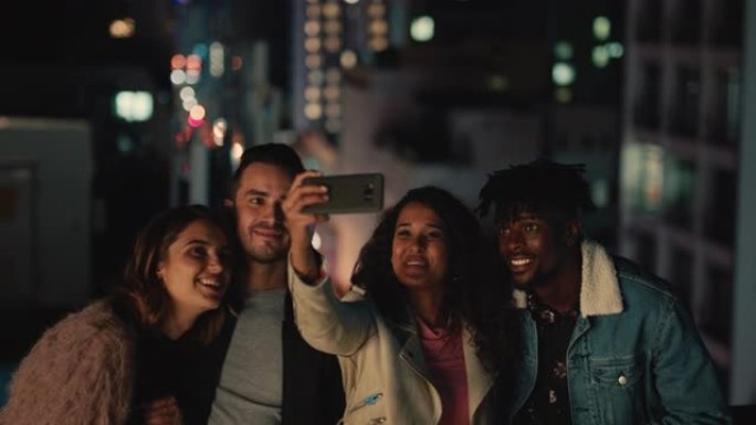年轻的多民族朋友晚上在屋顶上摆姿势合影庆祝友谊团聚年轻女子使用智能手机分享周末聚会到社交媒体