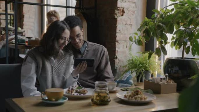 女孩和男孩学生的慢动作肖像在智能手机屏幕上观看有趣的视频在咖啡馆的餐桌上笑