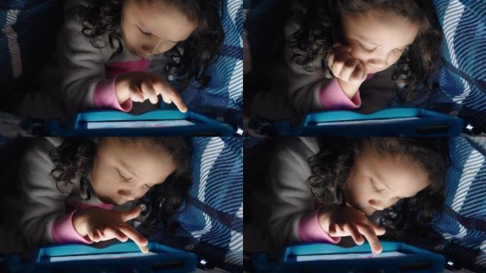 可爱的小女孩在毯子下使用数字平板电脑享受触摸屏技术玩游戏睡觉时玩得开心