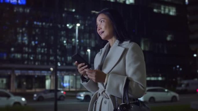 中年亚洲妇女的肖像晚上在城市街道上行走，使用智能手机。优雅的女商人微笑，发短信，在社交媒体上聊天，通