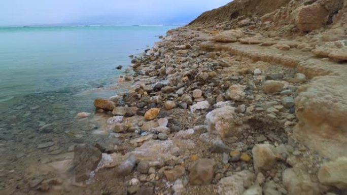 在死海沿岸行走，沙子和石头上覆盖着结晶盐，清澈的水接近以色列Ein Bokek海滩的典型风景