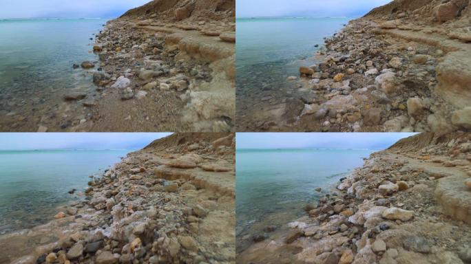在死海沿岸行走，沙子和石头上覆盖着结晶盐，清澈的水接近以色列Ein Bokek海滩的典型风景