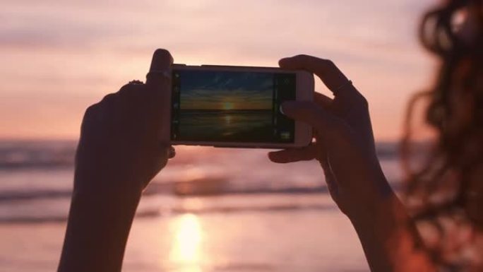 关闭海滩上的年轻女子使用智能手机拍摄美丽的日落享受分享暑假旅游经验在社交meda