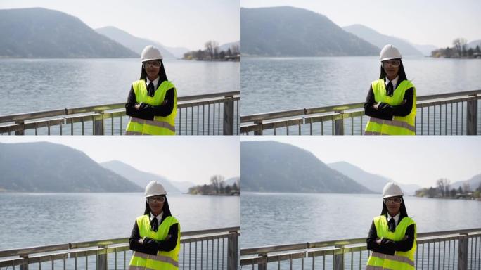 女工程师站在背心，防护眼镜和安全帽头盔上，对着镜头微笑，可以看到后面的湖