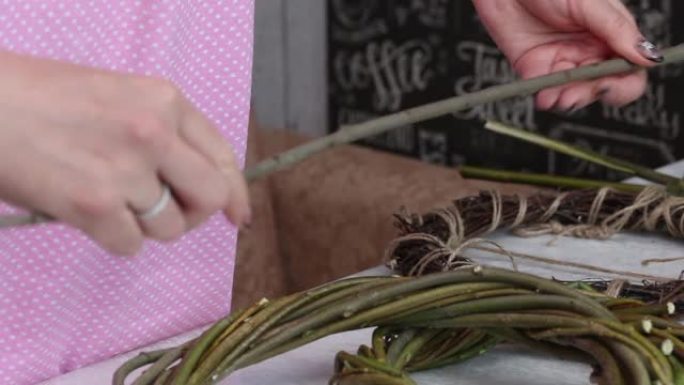 一个女人手里拿着藤蔓的树枝来编织。藤蔓编织的花环躺在桌子上。不同的尺寸。附近的藤蔓树枝。特写。