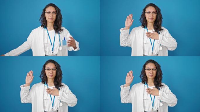 年轻美丽的西班牙裔女医生用手在孤立的蓝色墙壁背景上宣誓