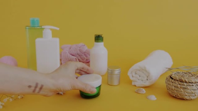 女性手把身体护理霜放在瓶子旁边，黄色背景上有有机化妆品和浴室配件