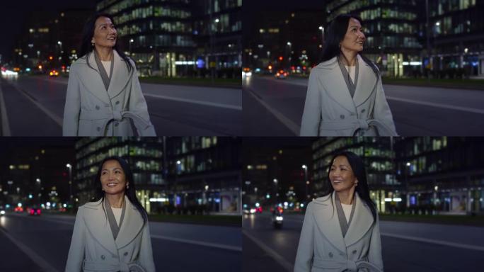 真实的中年亚洲妇女的肖像晚上走在城市街道上，欣赏风景。优雅的女导演出差微笑，享受城市生活和城市灯光