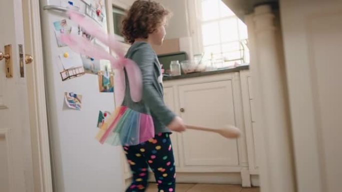 快乐的小女孩戴着可爱的仙女翅膀在厨房跳舞，挥舞着勺子玩得开心，假装做有趣的舞蹈动作，享受周末在家