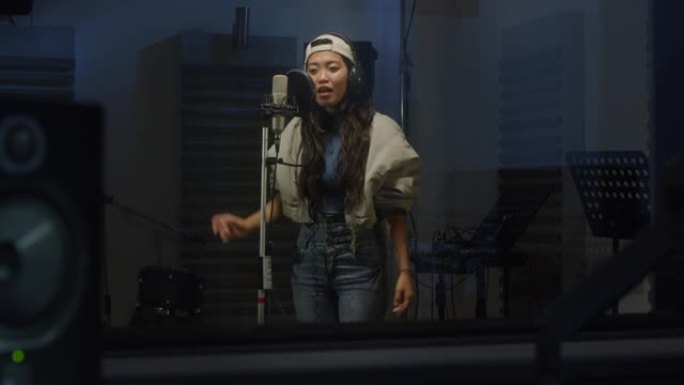 年轻的亚洲女歌手肖像，戴着耳机在专业录音室录制专辑。富有表现力的年轻女子带着情感唱歌，与广播听众分享