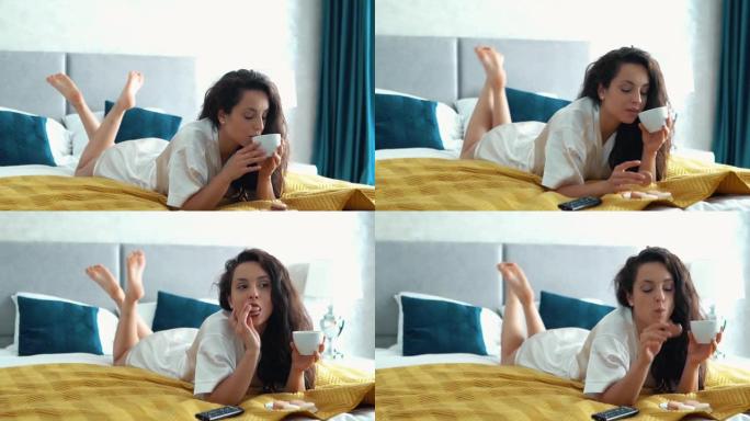 性感美丽的白人女孩躺在她的床上在她的卧室，看电视感兴趣，喝咖啡从白色的杯子和吃饼干。一个女人在吃巧克