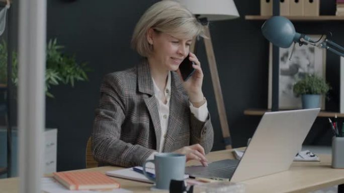开朗的女商人在手机上讲话并在办公室室内使用笔记本电脑的肖像