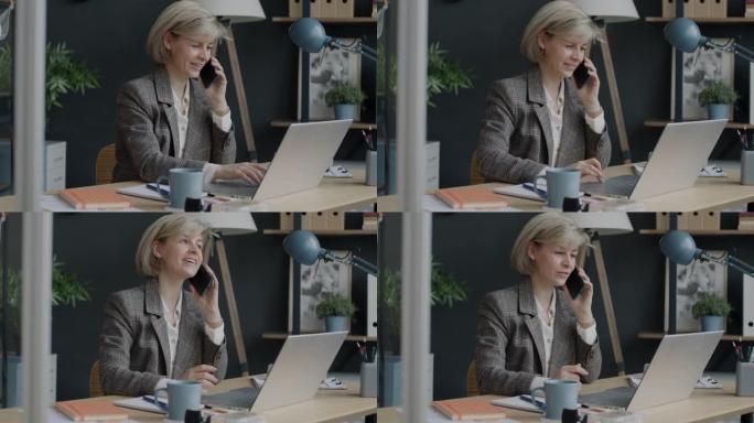 开朗的女商人在手机上讲话并在办公室室内使用笔记本电脑的肖像