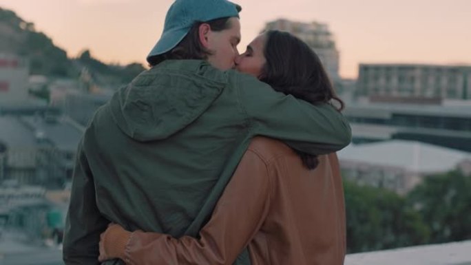 年轻的高加索夫妇在屋顶上接吻，拥抱浪漫，分享亲密的联系，在日落时享受城市的景色