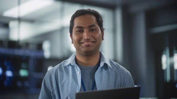 英俊的微笑的IT专家在数据中心使用笔记本电脑，看着相机。软件开发机构中成功的南亚商人和电子商务企业家