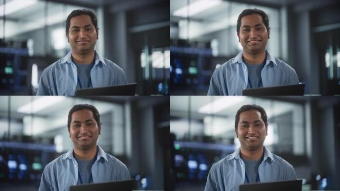 英俊的微笑的IT专家在数据中心使用笔记本电脑，看着相机。软件开发机构中成功的南亚商人和电子商务企业家