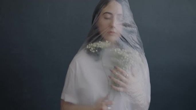 包裹着塑料的年轻女子的肖像，拿着鲜花站在灰色背景下