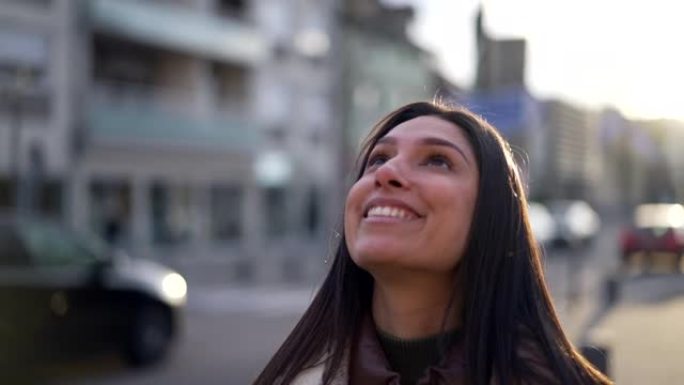 一位充满希望和信念的年轻女子站在街上看着天空。20多岁的快乐女性站在城市背景特写脸部肖像