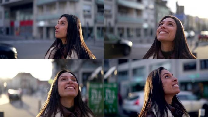 一位充满希望和信念的年轻女子站在街上看着天空。20多岁的快乐女性站在城市背景特写脸部肖像