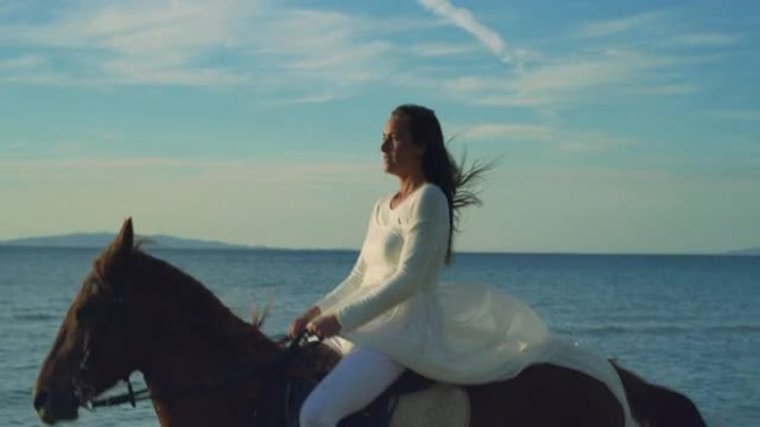 一个穿着白色衣服的美丽幸福女人的肖像骑着奔腾的马在海边，背景是明亮的蓝天。女性马术享受自由和清新的微