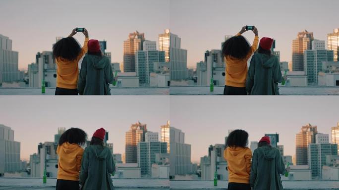 后视图年轻女性朋友日落时在屋顶上自拍，享受一起喝酒分享友谊，看着城市天际线