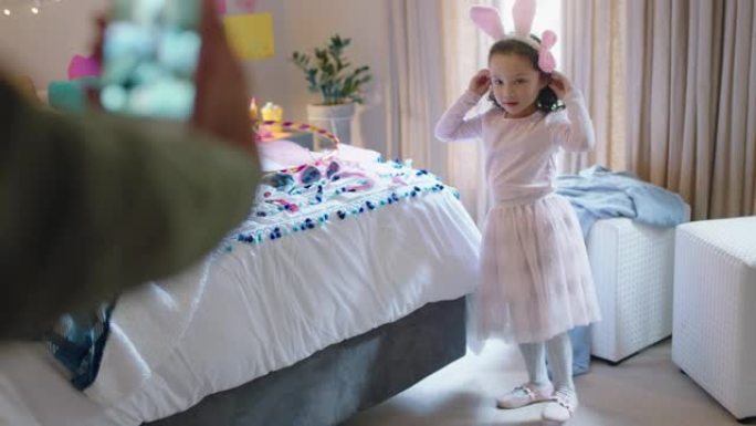 快乐的小女孩在卧室里穿着兔子耳朵玩耍，母亲在社交媒体上使用智能手机分享拍照