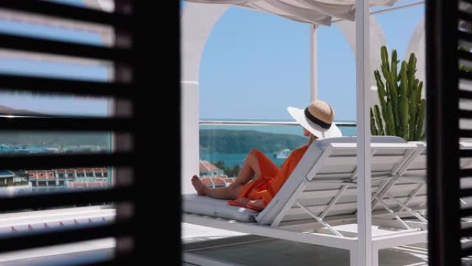 女人在日光浴床放松，在豪华屋顶顶楼露台享受暑假，可以看到海景。后视图
