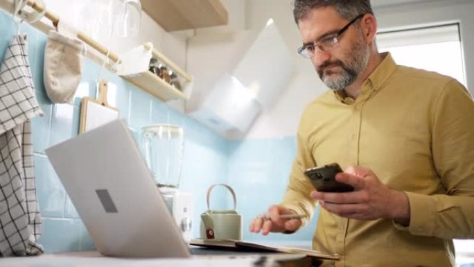 男人在现代厨房的笔记本电脑上工作时使用手机
