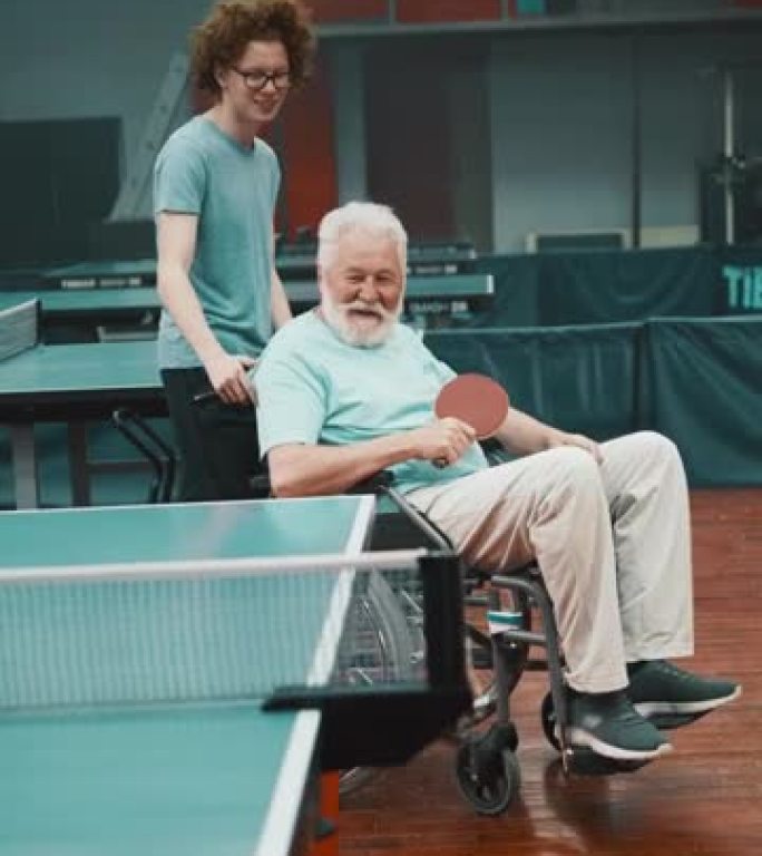 一个十几岁的少年带他开朗的祖父在俱乐部打乒乓球，推着他坐在轮椅上的垂直镜头