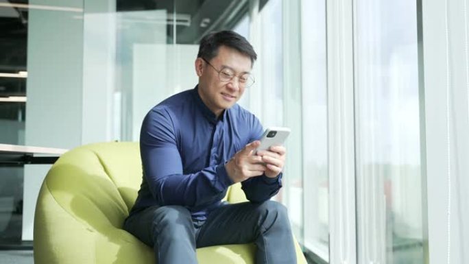 亚洲商人IT程序员坐在豆袋椅上，拿着智能手机滚动，在现代办公室工作场所观看社交媒体