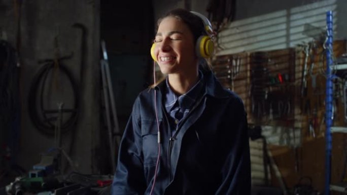 穿着制服工作服的年轻女机械师的肖像戴上音乐耳机，听着快乐的音乐。在维护车间休息的焊工职业女性