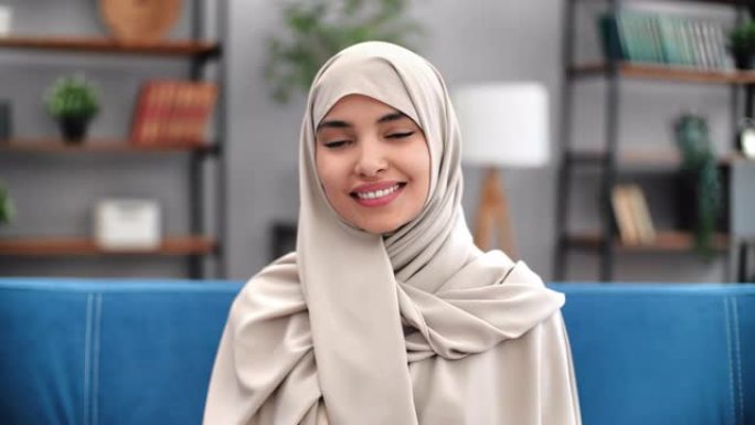 肖像年轻穆斯林妇女米色头巾在现代家庭室内微笑在沙发上摆姿势