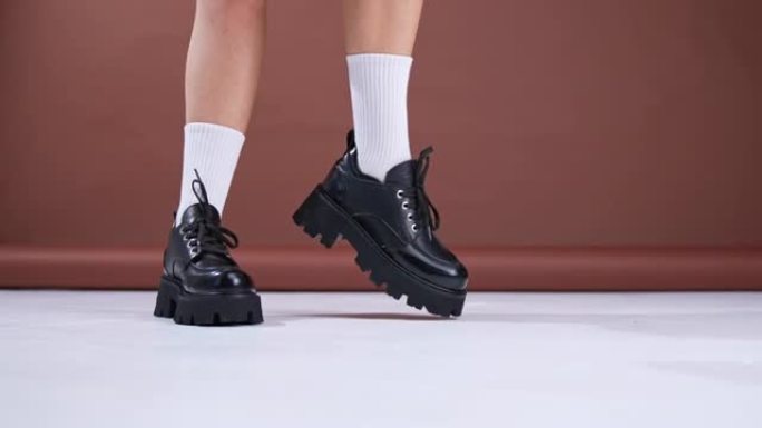 大型拖拉机鞋底上的黑色皮革秋鞋。穿着白色袜子的模特展示了她在镜头前跳舞的现代时尚鞋类。特写。