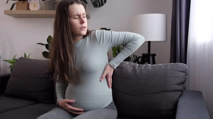 心烦意乱的年轻孕妇坐在家里的沙发上按摩背部肌肉，患有严重的疼痛感觉，未来的妈妈在妊娠晚期大肚子下背部